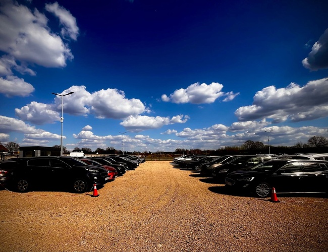 GoPark – Aparcamiento de gravilla lleno de coches bajo el cielo azul. Aquí están aparcados coches alineados. Todo está iluminado por altas lámparas.