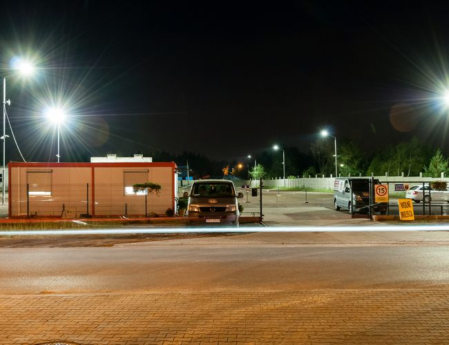 AAP-66 – Oświetlony parking z kontenerem biurowym. Widać zaparkowane samochody i płot. Tablica informuje o wolnych miejscach.