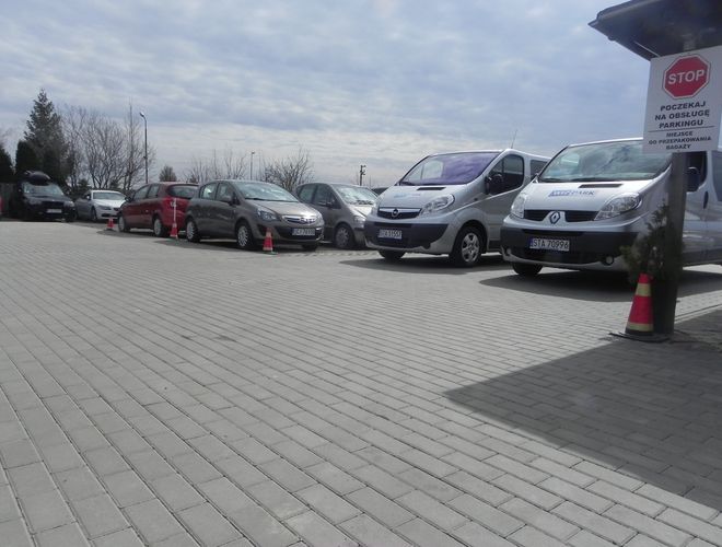 WIZ-PARK – Parkujące samochody osobowe i dostawcze stoją w rzędzie. Parking wyłożony jest kostką brukową. Obok stoi znak stopu.