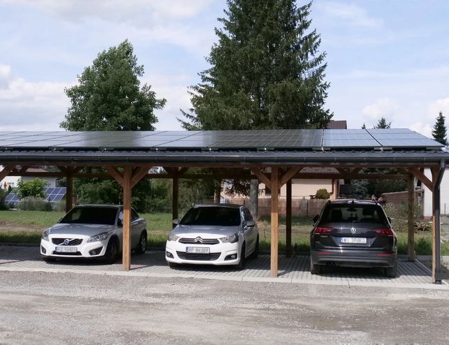 Parking Zielony Kompleks Dwór Ostoya - trzy samochody pod zadaszeniem z panelami słonecznymi. Teren częściowo otoczony drzewami.