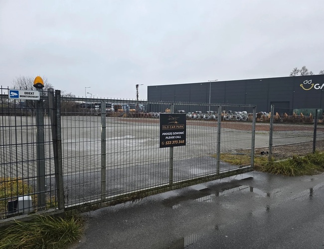 Parkovisko Olo Car Park - Oplotený priestor s kovovou bránou. Miesto je pod dohľadom. Na pozadí je vidieť veľkú budovu.