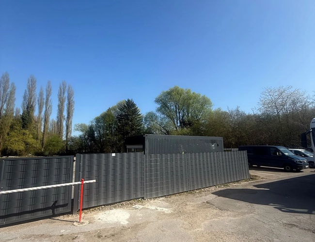 Parking Trust Park Fly - Ogrodzony teren z szarym płotem. Obok zaparkowane są czarne furgonetki. W tle widać drzewa.