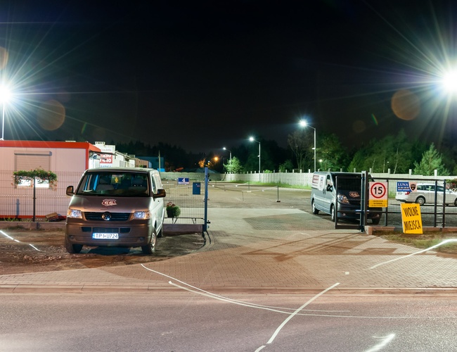 AAP-66 – Parking ogrodzony, dobrze oświetlony. Wjazd z bramą. Dwa zaparkowane pojazdy, wolne miejsca dostępne.