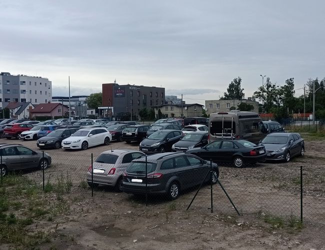 Smart parking - Na nieogrodzonym terenie stoi wiele samochodów. W tle widać budynki i drzewa. Obok są przenośne toalety i kontener.