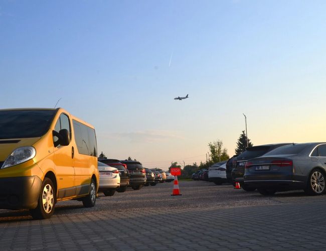 Royal Parking – Parking pełen samochodów. Nad nim leci samolot. Słupki ostrzegawcze stoją na placu.