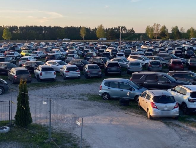 Parkolás a széltorony alatt - A nagy téren számos autó áll. A terület bekerített és nyitott. Háttérben fák láthatók.