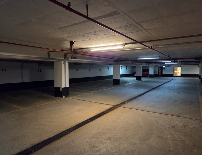 Atomis Hotel Munich Airport Parking Cubierto – Estacionamiento subterráneo con espacios numerados. Techo iluminado y coche visible en la esquina.