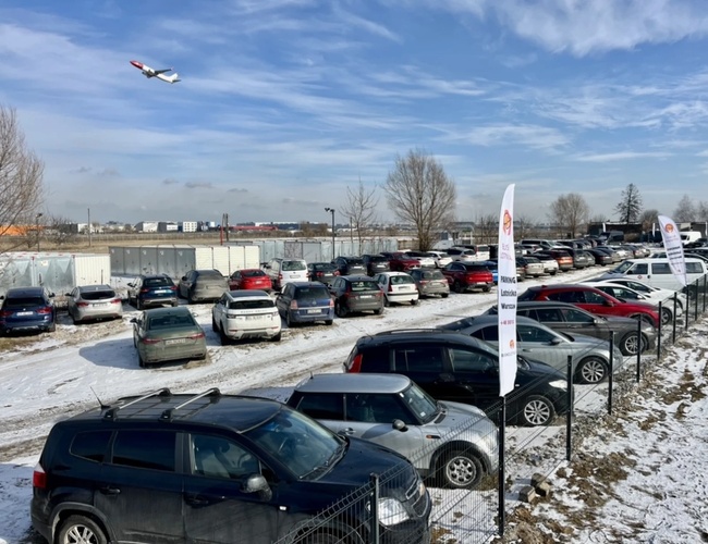 Parkovanie blízko letiska - ohradený areál s zaparkovanými autami. Na oblohe lieta lietadlo. Zimná krajina a modré nebo.