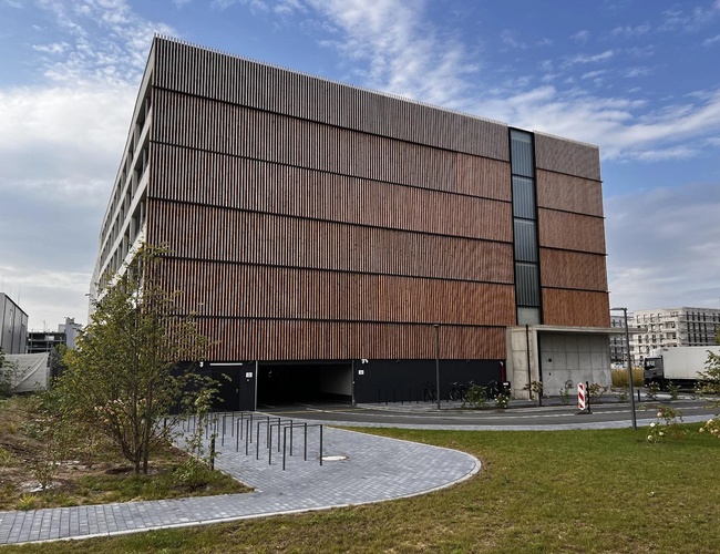 Parkovisko na letisku Paringový sluha – Vysoká, viestupňová budova s drevenou fasádou. Pred ňou je cesta a trávnik.