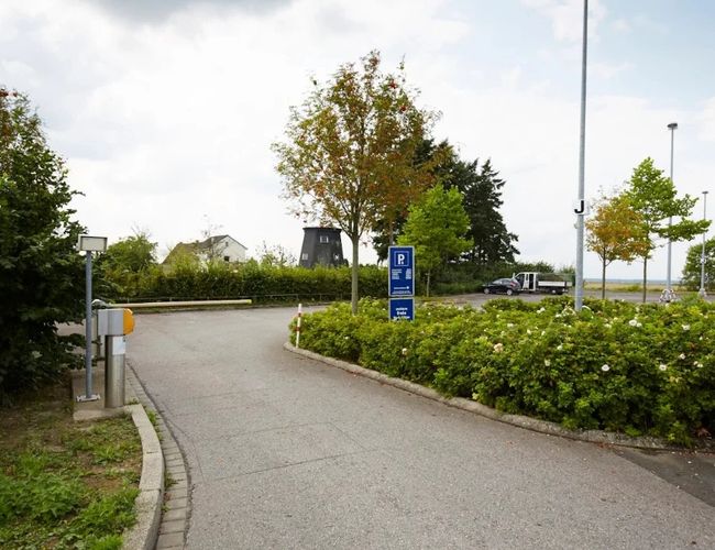 Droga dojazdowa do parkingu z oznakowaniem przy wjeździe. Po obu stronach drogi znajdują się krzewy i drzewa, a w tle widoczny jest teren parkingu.