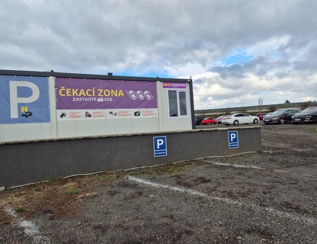 HostParking - Na zdjęciu widoczny jest kontener biurowy obok zaparkowanych samochodów. Teren parkingu jest nieutwardzony i oznakowany.