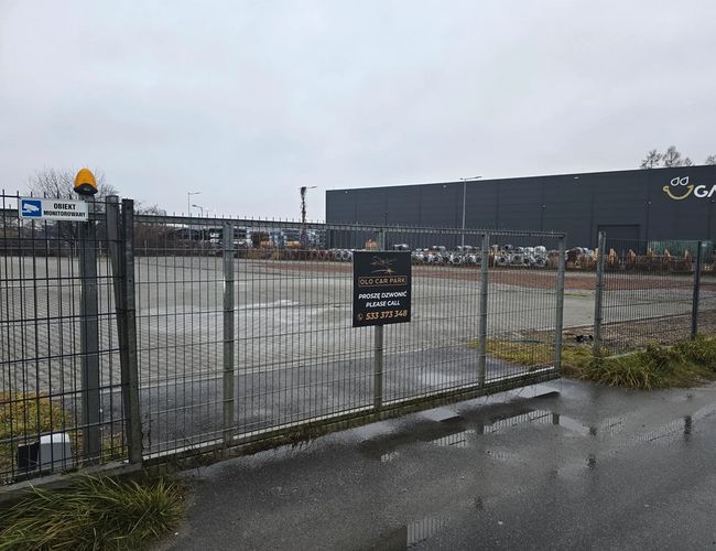 Parking Olo Car Park - Ogrodzony teren z metalową bramą. Miejsce jest monitorowane. W tle widoczny duży budynek.