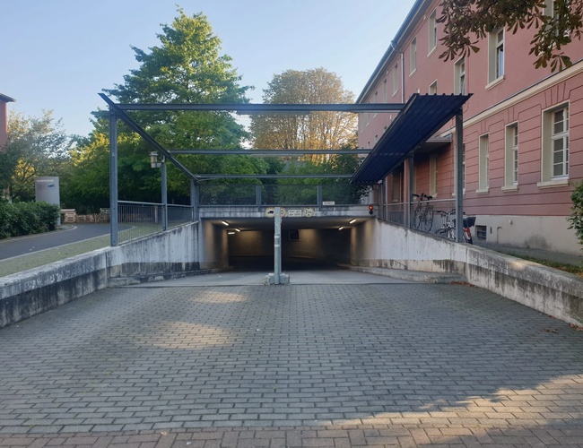 Wejście do podziemnego parkingu w Frankfurcie z rampą w dół, otoczone zielenią i budynkiem w tle.