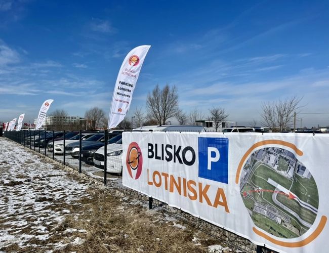 Parking Blisko Lotniska - Ogrodzony teren z zaparkowanymi samochodami. Flagi reklamowe wzdłuż ogrodzenia. Zimowy krajobraz w tle.