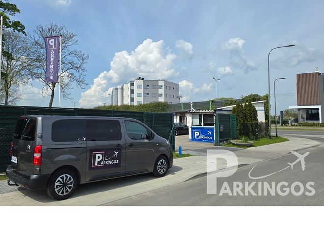 Parking Parkingos - Widoczny ogrodzony teren z autem dostawczym. Obok stoi mały budynek. Na trawie rosną tuje.