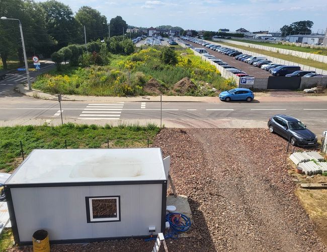Sky Park - Duży parking z wieloma zaparkowanymi samochodami. Widać kontener na terenie. Obok krzewy i skrzyżowanie z pasami.