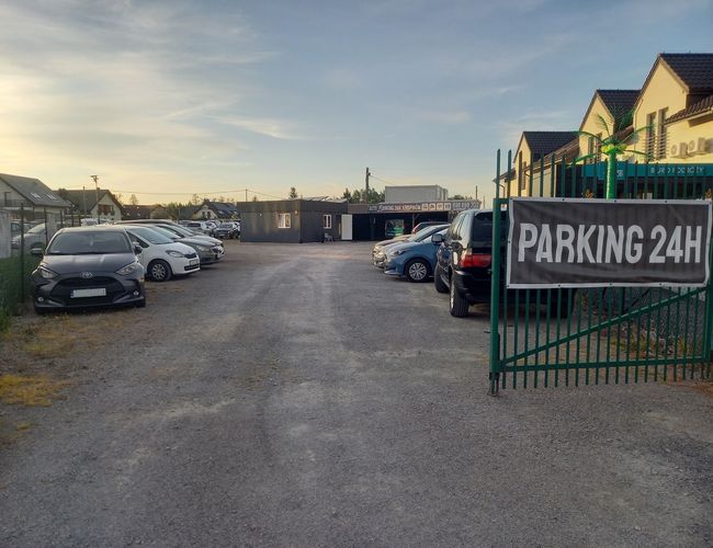 AutoParking24h - Parking jest żwirowy i ogrodzony. Samochody stoją w dwóch rzędach. Obok jest budynek obsługi.