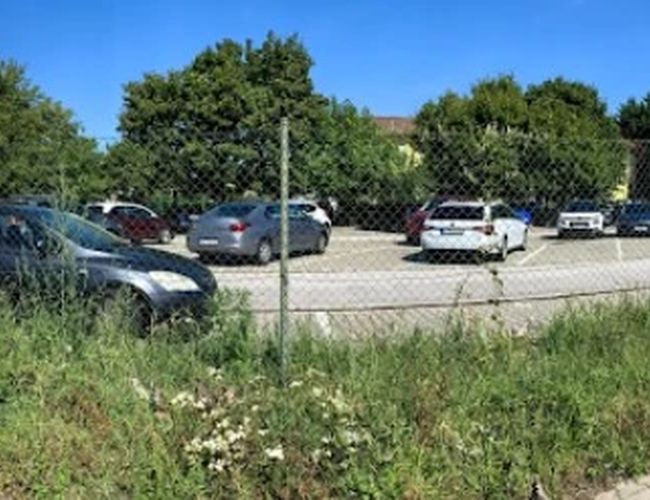 Ogrodzony parking z zaparkowanymi samochodami i budynkiem w tle, otoczony zielenią.