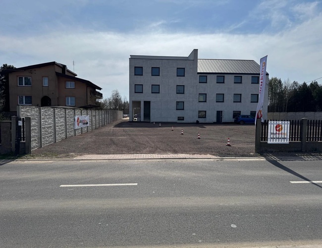 SkyParking - Plaza pavimentada con lugares para coches. El aparcamiento está cercado y tiene postes de carretera. Detrás se encuentra un edificio de oficinas.