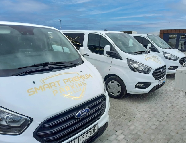 Smart Premium Parking - Na parkingu stoją cztery białe busy. Każdy ma logo Smart Premium Parking na masce. Pojazdy są zaparkowane na kostce brukowej.