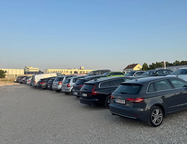 Rząd samochodów zaparkowanych na parkingu. Widoczna utwardzona nawierzchnia i budynki w tle.