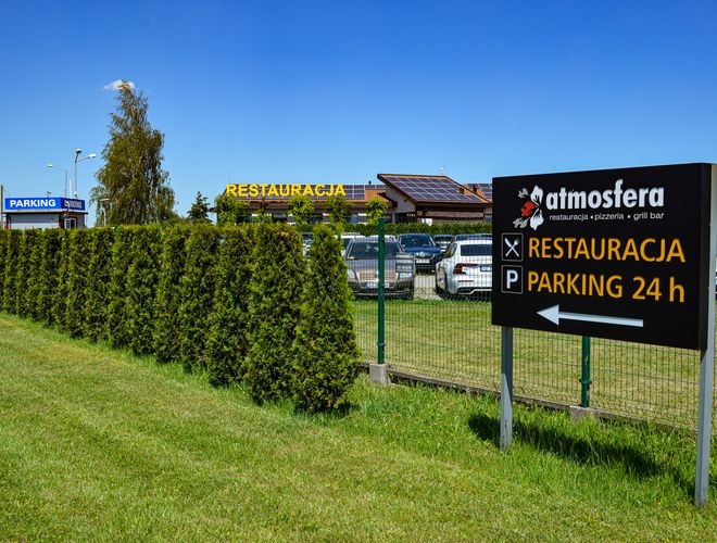 Parking Autosfera - Widoczny znak informuje o restauracji i parkingu. Ogrodzenie z żywopłotem oddziela strefę parkowania.