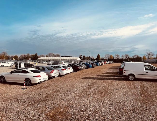 R8 Parking – Na żwirowym placu zaparkowane są samochody. Parking jest pełen aut różnych marek. W tle widać drzewa i budynek.
