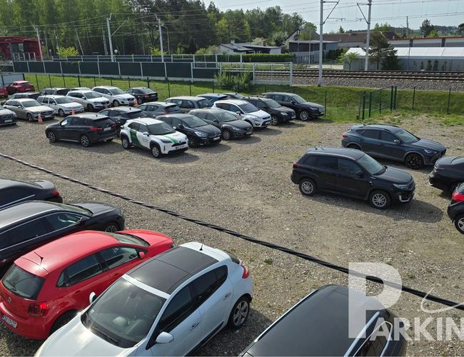 Parking Parkingos - Teren jest nieutwardzony i otoczony ogrodzeniem. Samochody stoją zaparkowane w rzędach. W tle widać zielone drzewa i tory kolejowe.