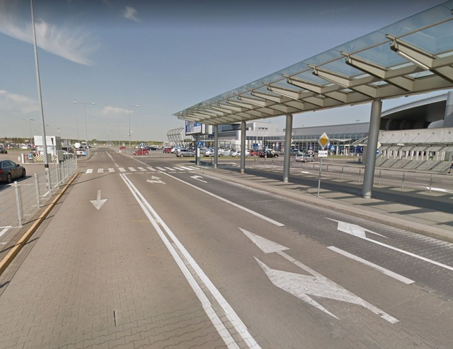 Oficiálne parkovisko P2 - Cesta vedie pozdĺž zastrešenej chodby. Vedľa sú zaparkované autá a budova s nápismi.