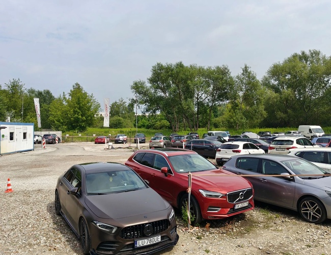 WAW Parking - Linii lungi de mașini parcate pe o suprafață de pietriș. Teren împrejmuit cu copaci pe laturi.