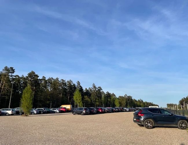 Auto Airport Parking - Parking jest żwirowy i otoczony drzewami. Samochody stoją w rzędach. Pogoda jest słoneczna.