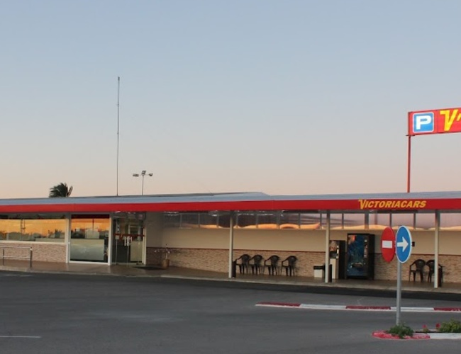 Victoria Parking Alicante – Edificio con techo rojo y logo. Al lado hay una furgoneta blanca. En el fondo, cielo y palmeras.