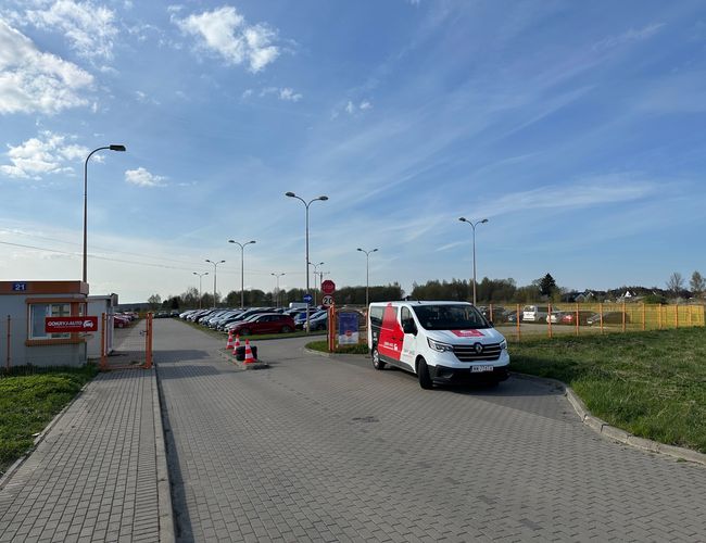 Odkryj-Parking GDN - Teren otoczony ogrodzeniem. Na parkingu stoją samochody. Obok przejeżdża biały van.