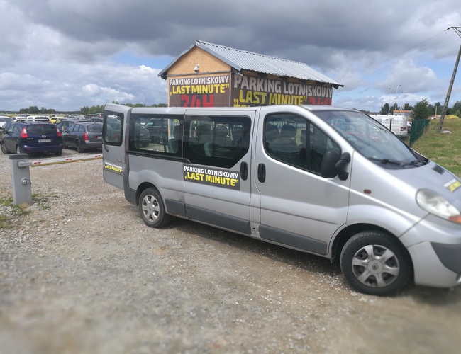Parking P8 - Na placu stoi bus z reklamą parkingu. W tle widać zaparkowane samochody. Teren jest żwirowy i ogrodzony.