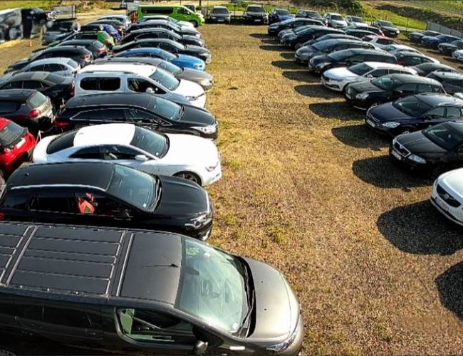 Gran Aparcamiento - Se pueden ver coches aparcados sobre una superficie de grava. El aparcamiento está lleno y el área está cercada.