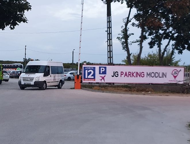 JG Parking Modlin - fehér minibuszos belépőkapu. A parkoló kerítéssel van körülvéve, és fákkal van körülötte.
