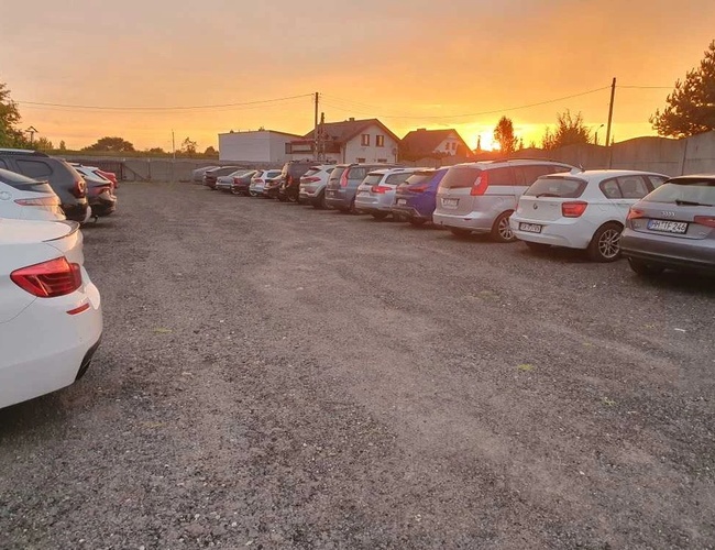 Parking Wings - En el estacionamiento de grava hay muchos coches. El estacionamiento está rodeado de una valla. En el horizonte, el atardecer.
