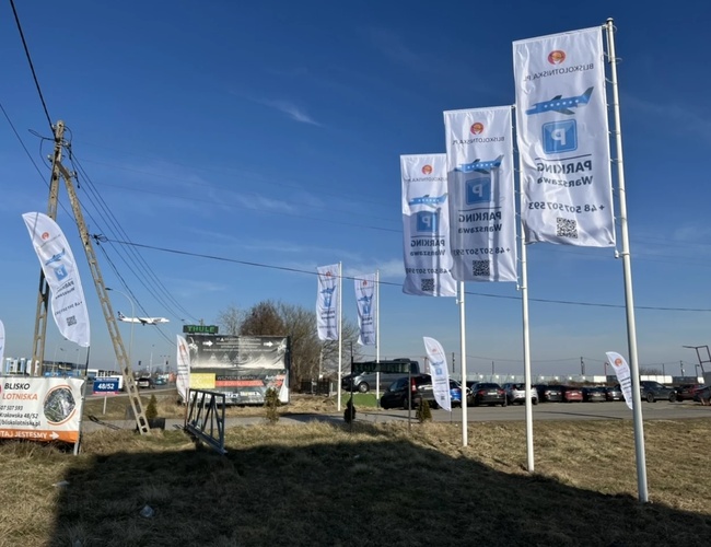 Parkovanie blízko letiska – viditeľné veľké vlajky a informačné tabule. Niekoľko zaparkovaných áut na spevnenom povrchu. Slnčný deň, čistá obloha.