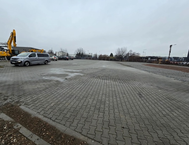 Parkovisko Olo Car Park – Na spevnenom nádvorí stoja autá a stavebné stroje. Pozemok je otvorený a priestranný. Okolo rastú stromy.