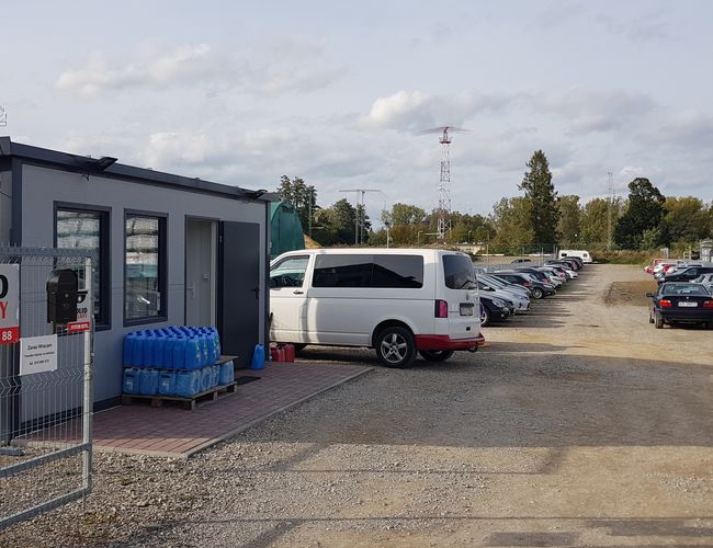 BaliceLot24 - Parking jest ogrodzony i chroniony. Stoi dużo samochodów. Obok znajduje się biały budynek.