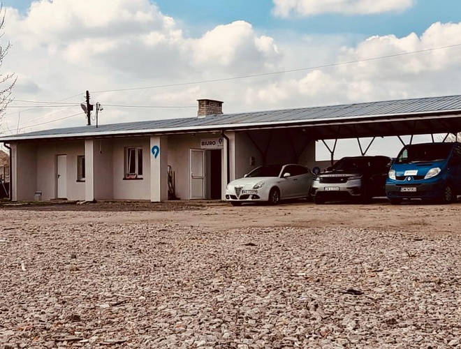 Parking MPark - Aparcamiento con base de grava y techado. Adyacente a él hay un edificio de oficinas. En el aparcamiento hay varios coches.