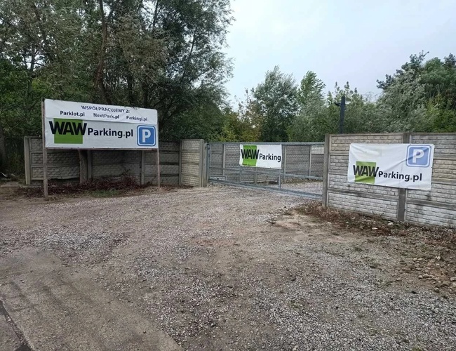 WAW Parking – Vstupná brána s reklamnými banermi. Kamenná plocha obklopená plotom. Stromy na pozadí.