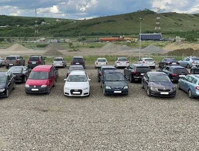 Parking pełen samochodów ustawionych na żwirowym placu, w tle zielone wzgórza i niebo z chmurami.