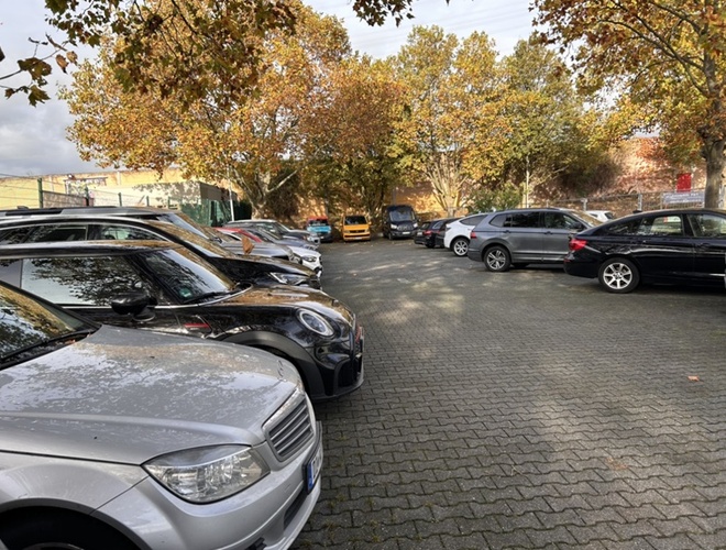 Parkplatz Parkinsel24 VALET - Autos stehen auf einem großen Platz. Im Hintergrund sind Bäume mit herbstlichen Blättern sichtbar. Der Platz ist geräumig und hat eine gepflasterte Oberfläche.