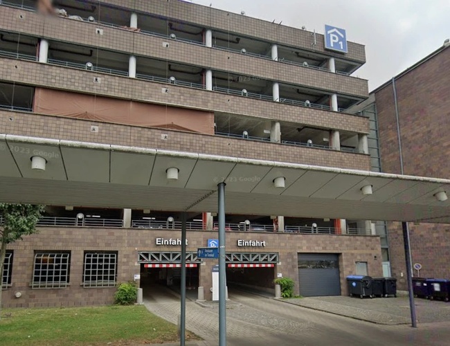 Widok na wielopoziomowy parking od strony wejścia. Przed budynkiem znajduje się zadaszenie nad strefą wejściową oraz oznakowanie parkingu.