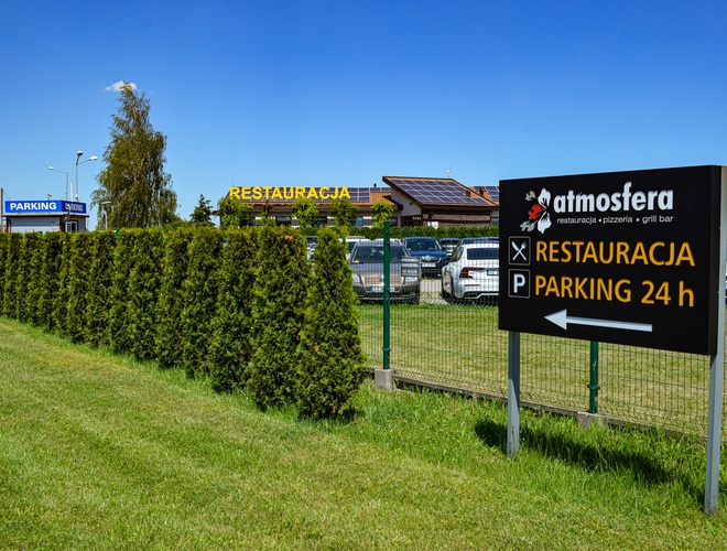 Parking Autosfera - Un letrero visible informa sobre el restaurante y el estacionamiento. Una cerca de arbustos separa la zona de estacionamiento.