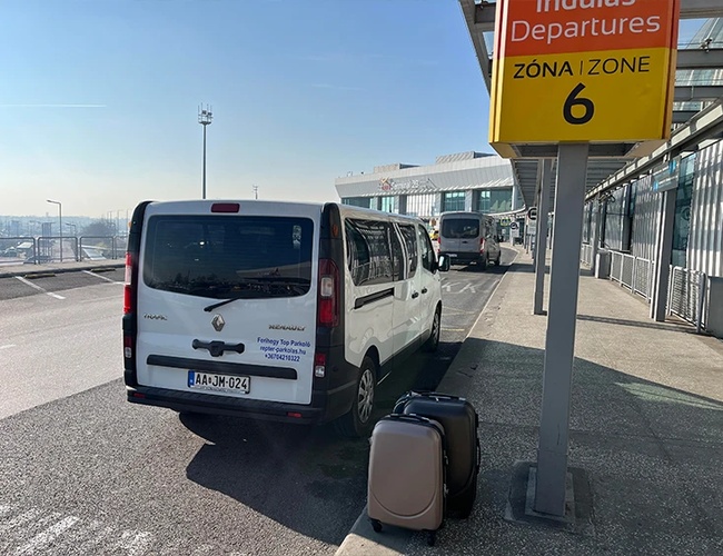 Bus stojący przy terminalu odlotów pod znakiem ‘Departures Zone 6’, obok leżą torby podróżnych.