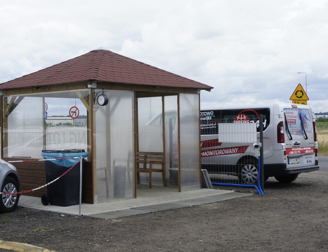 Parking Lotniskowy 24h - Obok altanki stoi samochód dostawczy. Obok leży kubeł na śmieci. W tle są tablice drogowe.