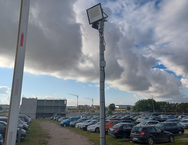 Goliat Parking — Widoczny duży, zatłoczony plac z samochodami. Teren jest nieutwardzony, z pojedynczym oświetleniem. W tle widać chmury i dźwigi.