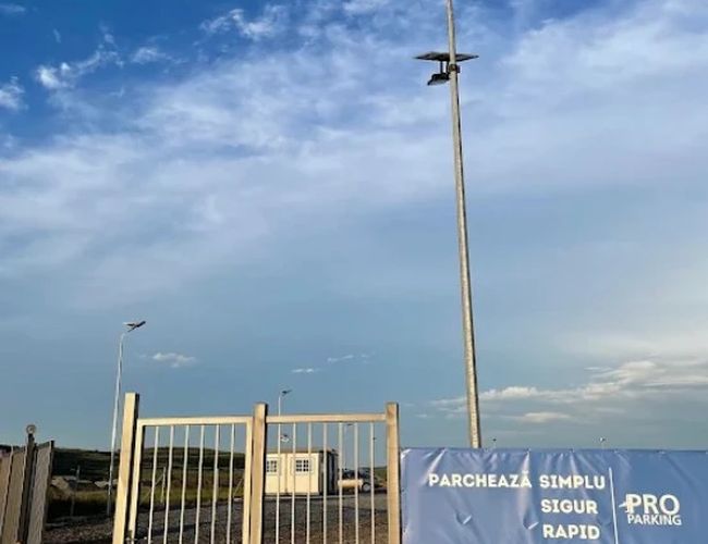 Brama wjazdowa z banerem informacyjnym i wysoką lampą, na tle nieba o zachodzie słońca.